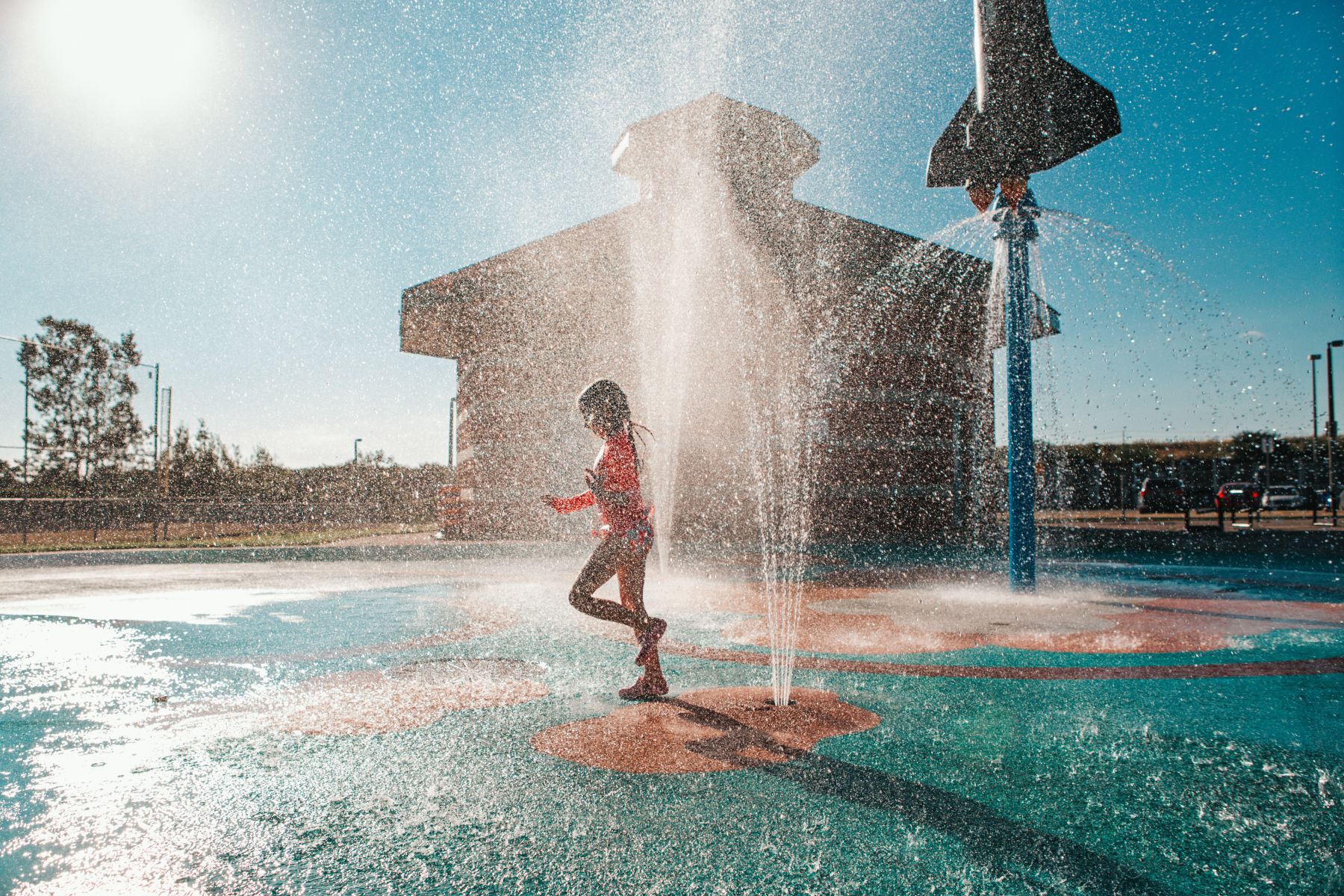 Child in a red swimsuit runs through a vertical water jet at a sunny splash pad with a rocket sculpture in the background.