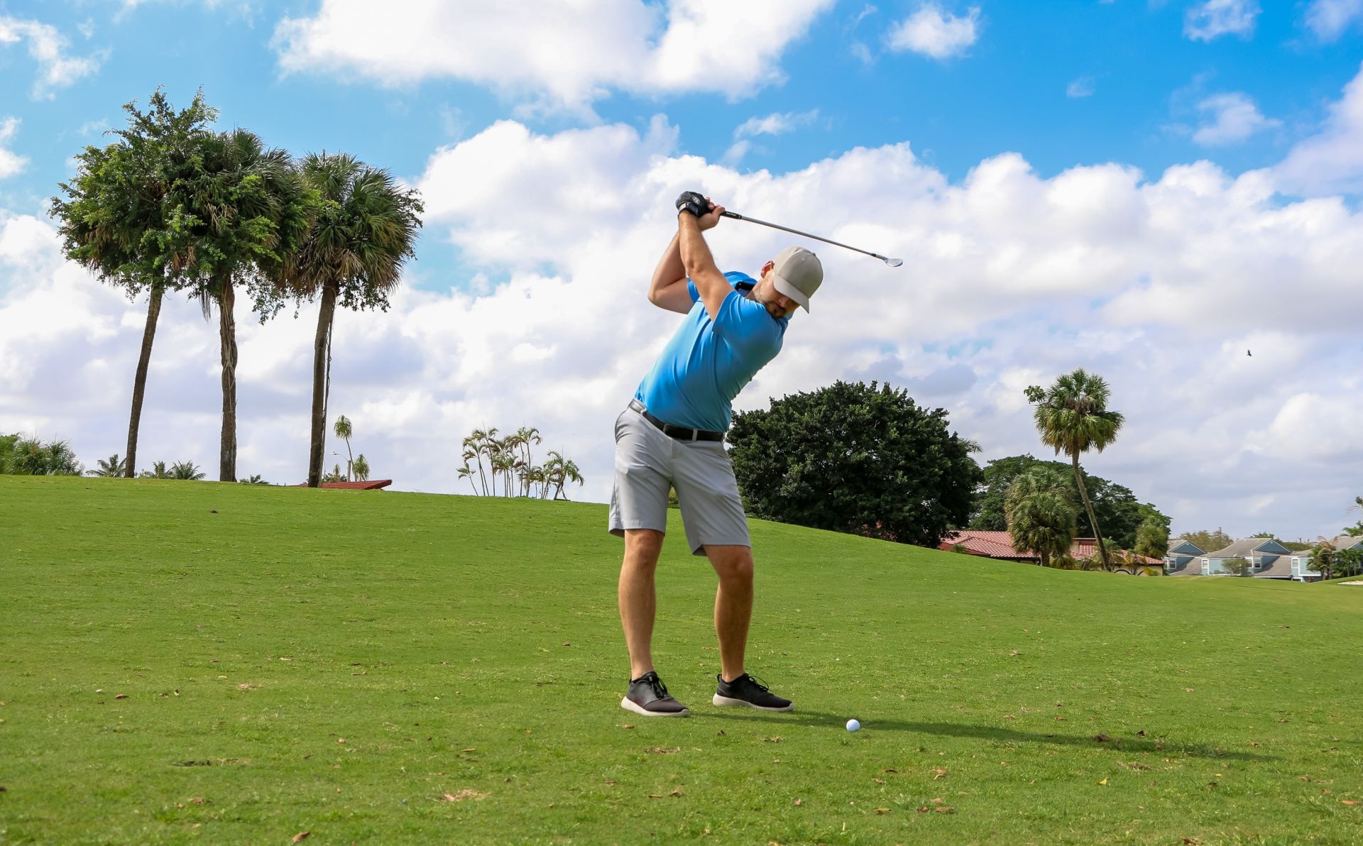 Man in a blue polo swings a golf club on a sunny course with palm trees and houses in the background.