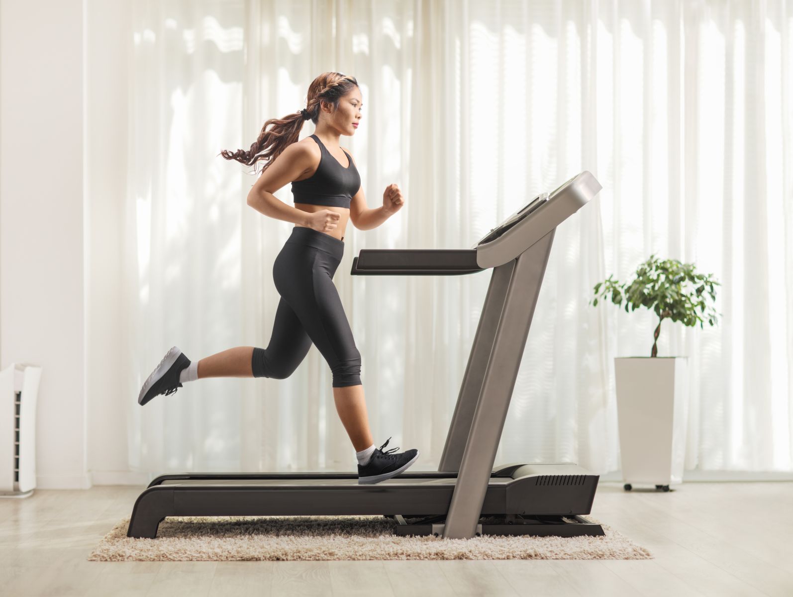 Woman in black athletic wear running on a treadmill in a bright home gym setting in a sunlit room.
